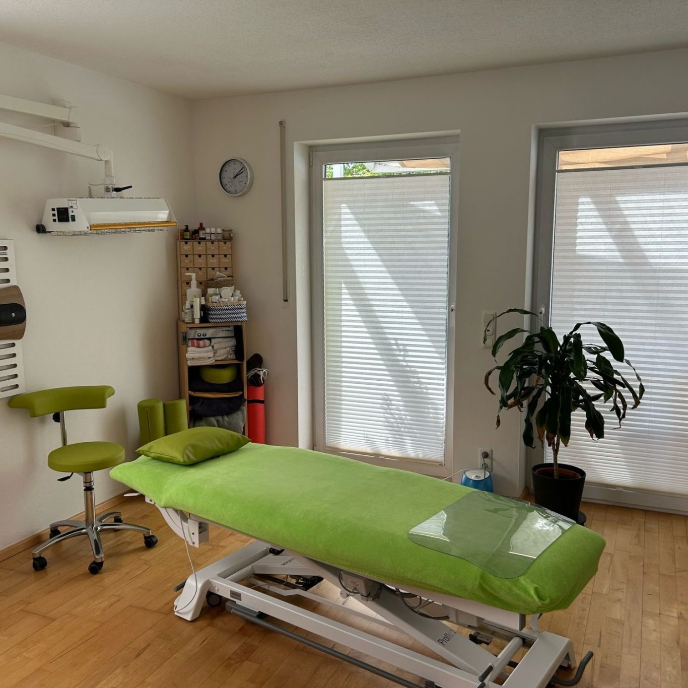 Behandlungsraum Behandlungsraum mit grünem Tisch, Stuhl und Pflanzen, großes Fenster mit Jalousien.